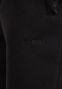 Schwarze Jogginghosen aus weichem Stoff verfügen über einen elastischen Bund und ein kleines erhabenes Logo "SMILODOX" an der Seite.