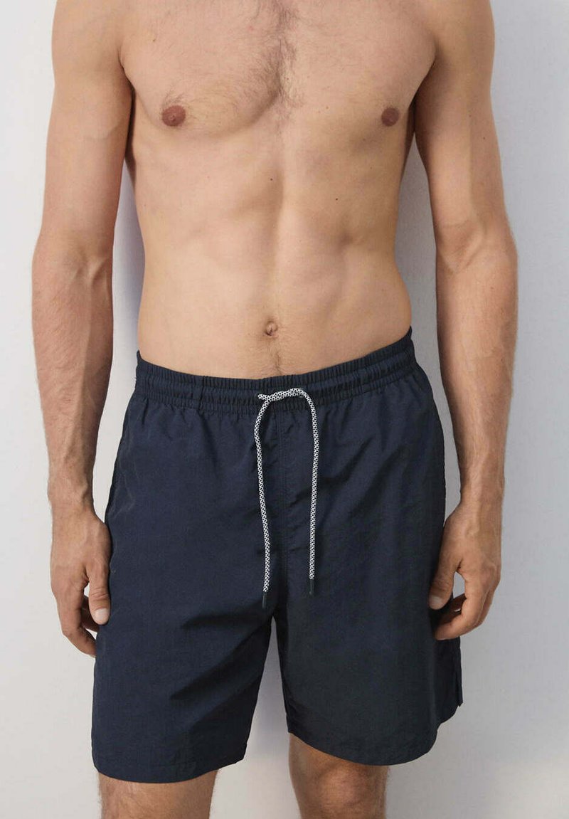 Homme portant un short de bain bleu marine foncé à cordon, visible de la moitié de la poitrine à la moitié de la cuisse sur fond clair uni.
