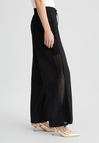 Pantaloni neri a gamba larga con pannelli laterali in rete e vita elastica. Dettaglio con cordino in vita. Tessuto testurizzato per la traspirabilità.