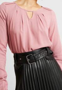Blusa a maniche lunghe rosa chiaro con scollatura a goccia e pieghe, abbinata a una gonna nera plissettata in eco-pelle e una cintura larga nera.