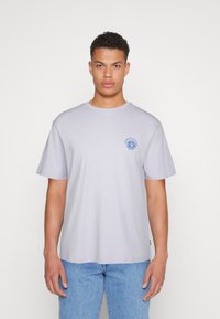 Tricou din bumbac gri deschis, cu mâneci scurte, de tip crew neck, având un grafic cu un soare albastru pe piept. Asortat cu blugi albaștri deschiși.