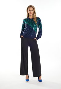 Navyblaues Paillettenoberteil mit grünen diagonalen Streifen, kombiniert mit schwarzen weiten Hosen. Das Model trägt blaue High Heels und hat die Hände in den Taschen.