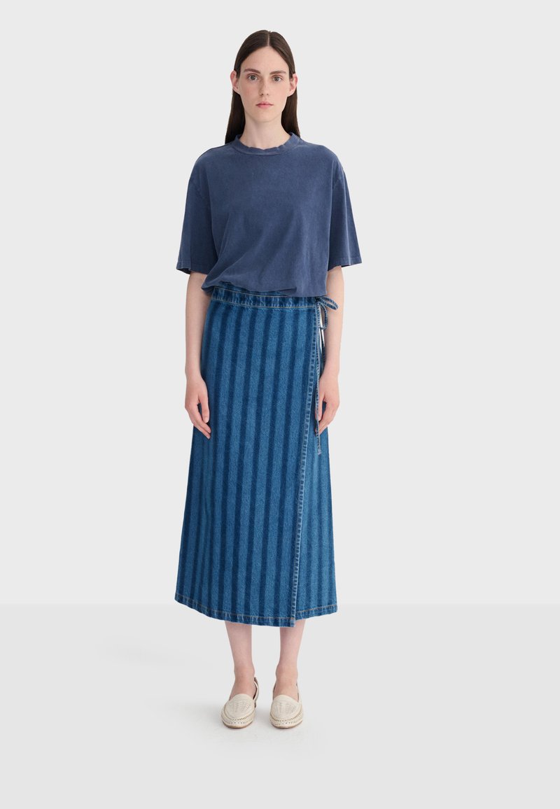 Femme aux longs cheveux bruns portant un t-shirt bleu ample, une jupe portefeuille en jean à rayures et des chaussures crème sans lacets, debout devant un fond uni.
