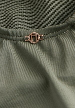 Kostkovaná látka olivově zelené barvy s žebrovanou strukturou, zdobená kulatou zlatou sponou s geometrickým výřezem.