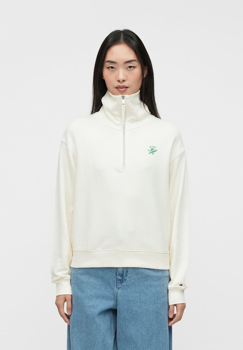 Ivory spalvos zip-up džemperis su aukštu apykakle, ant krūtinės su žaliai siuvinėta logo ir rausvomis rankovėmis. Minkšta, tekstūrinė medžiaga.