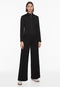 Schwarze kurze Jacke mit hohem Kragen und silbernem Reißverschluss, kombiniert mit weit geschnittenen schwarzen Hosen; glatte Textur, minimalistisches Design.