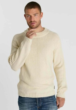 Uomo con capelli corti che indossa un maglione lavorato a maglia color crema e jeans blu, in piedi davanti a uno sfondo chiaro e uniforme, toccandosi il mento.