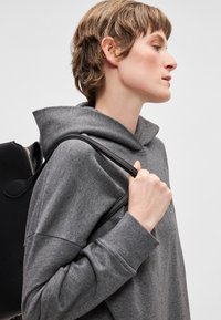 Šedá mikina s kapucí v pohodlném střihu, strukturovaná látka, a černý batoh se zipem, nošený přes rameno.