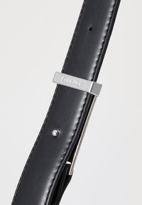 Fekete bőr öv sima textúrával, ezüstszínű fém csattal, amelyen a "Calvin Klein" logó van gravírozva. Egy kis fém szegecs díszíti.