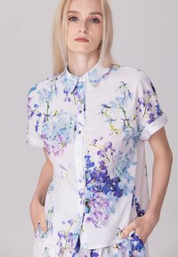 Chemise à manches courtes en tissu léger, ornée de motifs floraux bleus et violets sur fond blanc ; design à boutons sur le devant.