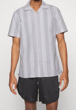 Homme portant une chemise à manches courtes boutonnée avec des rayures grises verticales et un short noir texturé, debout devant un fond uni.
