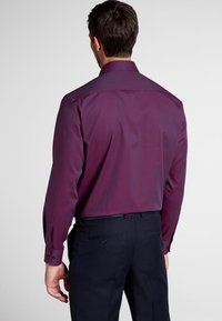 Koszula z długim rękawem, dopasowana, w głębokim purpurowym kolorze, o gładkiej fakturze, z usztywnionym kołnierzem i mankietami na guziki, zestawiona z ciemnymi spodniami.