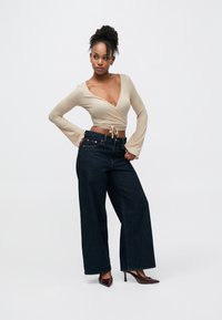 Bēša, ar garām piedurknēm, apkakles tops ar sasiešanas aizdari, kombinēts ar tumši zilām plaša piegriezuma džinsu biksēm. Modelis valkā bordo krāsas smailus papēžus.