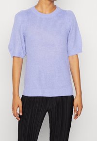 Frau in einem helllila gestrickten Kurzarmpullover mit Puffärmeln und schwarzen, vertikal gestreiften Hosen, die vor einem schlichten Hintergrund steht.