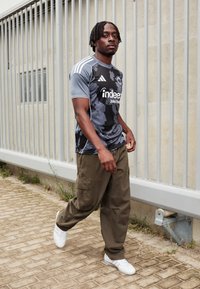 Jersey deportivo gris con patrón y acentos negros, logo blanco, pantalones cargo oliva y zapatillas blancas, caminando sobre una superficie pavimentada cerca de una valla.