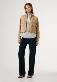 Tan suède korte jas met voorzakken en drukknoopsluitingen, gedragen over een licht geblokt shirt, gecombineerd met donker wijde jeans en beige hakken.