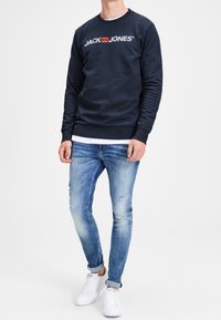 Uomo che indossa una felpa blu navy con il logo "JACK & JONES", jeans chiari arrotolati e sneakers bianche su uno sfondo bianco.