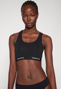 Soutien-gorge de sport noir raccourci avec un col arrondi, fabriqué en tissu lisse, comportant une large bandeau avec l'inscription 'PIECES' en blanc.