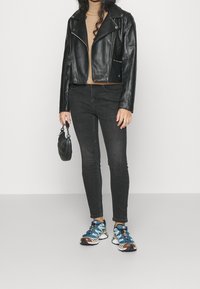 Veste de moto en cuir noir, pull beige, jean skinny noir, sac à main noir et chaussures de sport bleu et blanc avec des accents texturés.