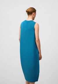 Ein ärmelloses, midi-langes blaues Kleid mit einem strukturierten diagonalen Faltenmuster. Der Stoff wirkt glatt und hat eine lockere Silhouette.