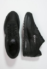 Pár černých tenisek Nike Air Max, jedna zobrazená z horního pohledu s tkaničkami, druhá z bočního pohledu, která zvýrazňuje vzduchové polstrování v podešvi a texturovaný design.