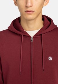 Bordeauxkleurige zip-up hoodie met een trekkoordcapuchon, voorzien van een klein wit logo op de borst. Zachte stof en platte stiksels zijn zichtbaar.