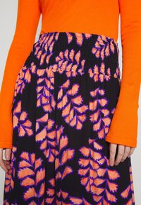 Langærmet orange top kombineret med sort skørt, der har lyse orange bladmønstre med lilla kanter og en smocked talje.