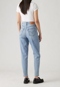 Kvinna som bär ljusblå högmidjade jeans med bakfickor och en vit kortärmad topp, som står framför en enfärgad bakgrund.