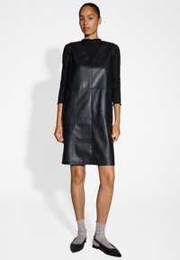 Zwarte leren A-lijn jurk met V-hals, voorzien van twee voorzakken en een gladde textuur, gecombineerd met doorschijnende sokken en zwarte ballet flats.