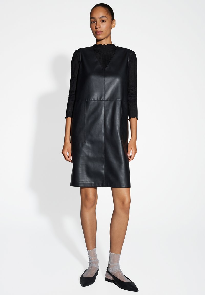 Zwarte leren A-lijn jurk met V-hals, voorzien van twee voorzakken en een gladde textuur, gecombineerd met doorschijnende sokken en zwarte ballet flats.
