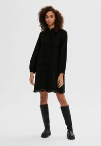 Schwarzes, knielanges Kleid mit rundem Ausschnitt, langen puffärmel und Spitzendetails. Zierliche Saumkante und Knopfverschluss vorne.