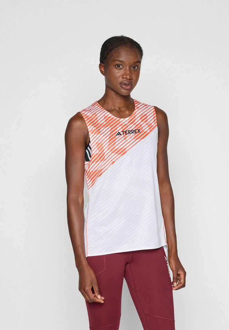 T-shirt de sport sans manches blanc avec des rayures diagonales orange, un motif texturé et le logo 'TERREX' en noir, associé à un legging bordeaux.