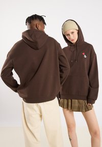 Bruine hoodie met een voorkeurszak, trekkoord en een kleine rode hartgrafiek. Vergezeld door een crème kleurige beanie en een geruite rok.