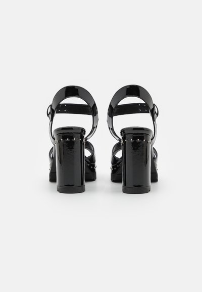 Sandalias de tacón alto de charol negro con tacones gruesos, correas ajustables en el tobillo y acentos de tachuelas. Se muestra la vista trasera.