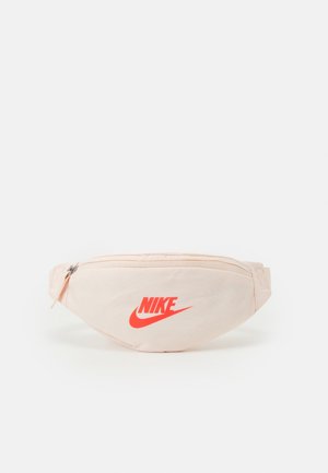 Beige Nike bæltetaske med rødt logo, lynlåslomme foran og justerbar rem på hvid baggrund.