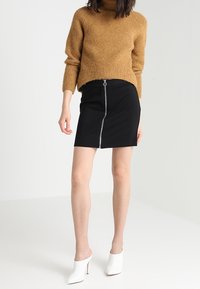 Hnědý žebrovaný svetr s vysokým límcem, doplněný černou mini sukní s předním zipem. Celý outfit doplňují bílé otevřené boty na podpatku.