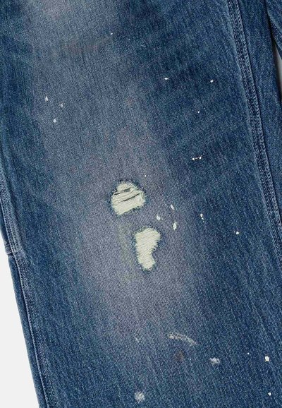 Jeans in denim blu con aree scucite e bordi sfrangiati, macchiati di vernice bianca. Il tessuto presenta un aspetto leggermente testurizzato e usurato.