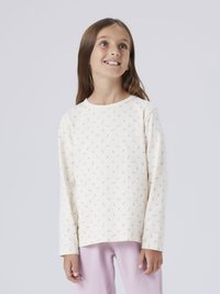 Fille aux longs cheveux bruns portant une chemise blanche à manches longues avec de petits imprimés floraux et un pantalon rose clair, regardant vers le haut et souriant.
