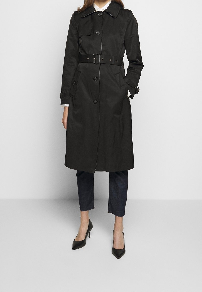 Trench-coat noir avec une taille cintrée par une ceinture, de gros boutons et un col pointu. Tissu lisse et résistant à l'eau, porté avec un jean foncé et des talons.