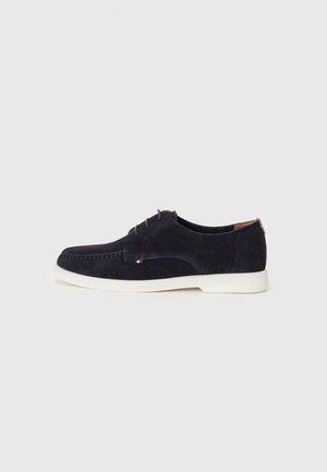 Chaussure décontractée en daim bleu foncé à lacets, semelle blanche et petit détail drapeau rouge, blanc et bleu sur le côté.
