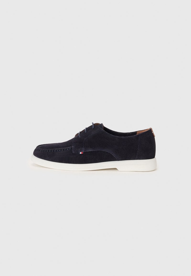 Chaussure décontractée en daim bleu foncé à lacets, semelle blanche et petit détail drapeau rouge, blanc et bleu sur le côté.