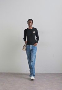 Egy ember magabiztosan sétál egy fekete pulóverben, amelyen bross van, kék farmerben és fehér sportcipőben, egy kis táskát hordozva, minimalista háttér előtt.