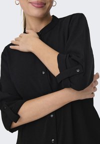 Camicia nera con colletto, tessuto testurizzato, maniche arrotolate e bottoni rotondi neri. La camicia ha una vestibilità ampia intorno alle spalle.