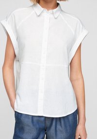 Camicia bianca in lino a maniche corte con bottone frontale, caratterizzata da un colletto a punta e una vestibilità rilassata, abbinata a pantaloni in denim blu.