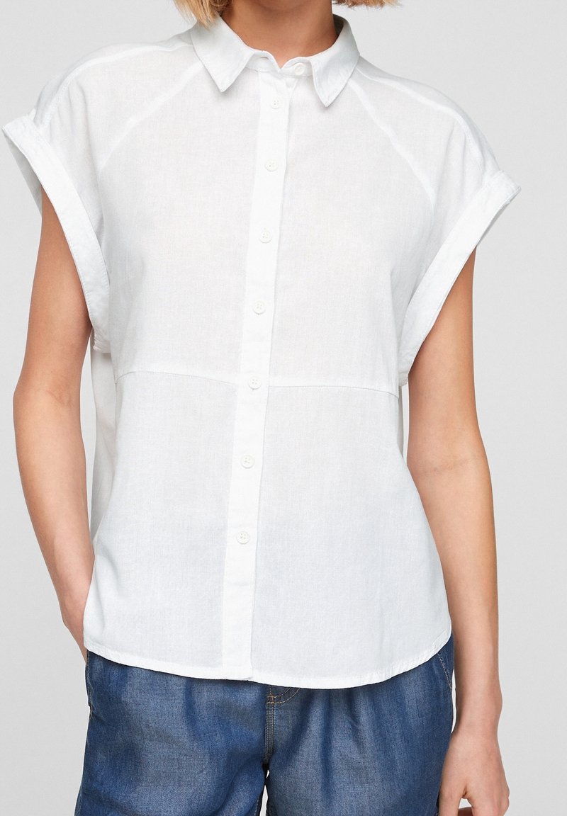 Camicia bianca in lino a maniche corte con bottone frontale, caratterizzata da un colletto a punta e una vestibilità rilassata, abbinata a pantaloni in denim blu.