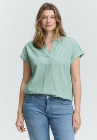 Light green short-sleeve top with a V-neckline, featuring a textured pattern. Paired with blue jeans, showing a relaxed fit. Textured fabric.