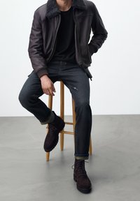 Mörkbrun läderjacka med shearling-krage, svart t-shirt, mörka denimjeans och mörkbruna snörkängor på en träpall.