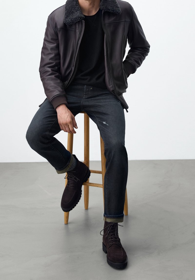 Mörkbrun läderjacka med shearling-krage, svart t-shirt, mörka denimjeans och mörkbruna snörkängor på en träpall.