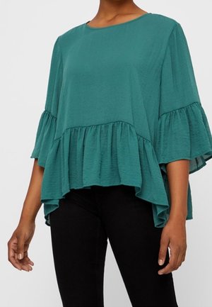 Blusa corta a maniche corte di colore teal con un design a strati e volant, texture morbida e scollatura rotonda. Abbinata a pantaloni neri.