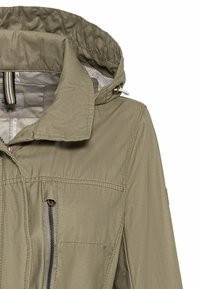 Veste vert olive clair avec une capuche, poche poitrine zippée et coutures matelassées sur le col et la doublure de la capuche.